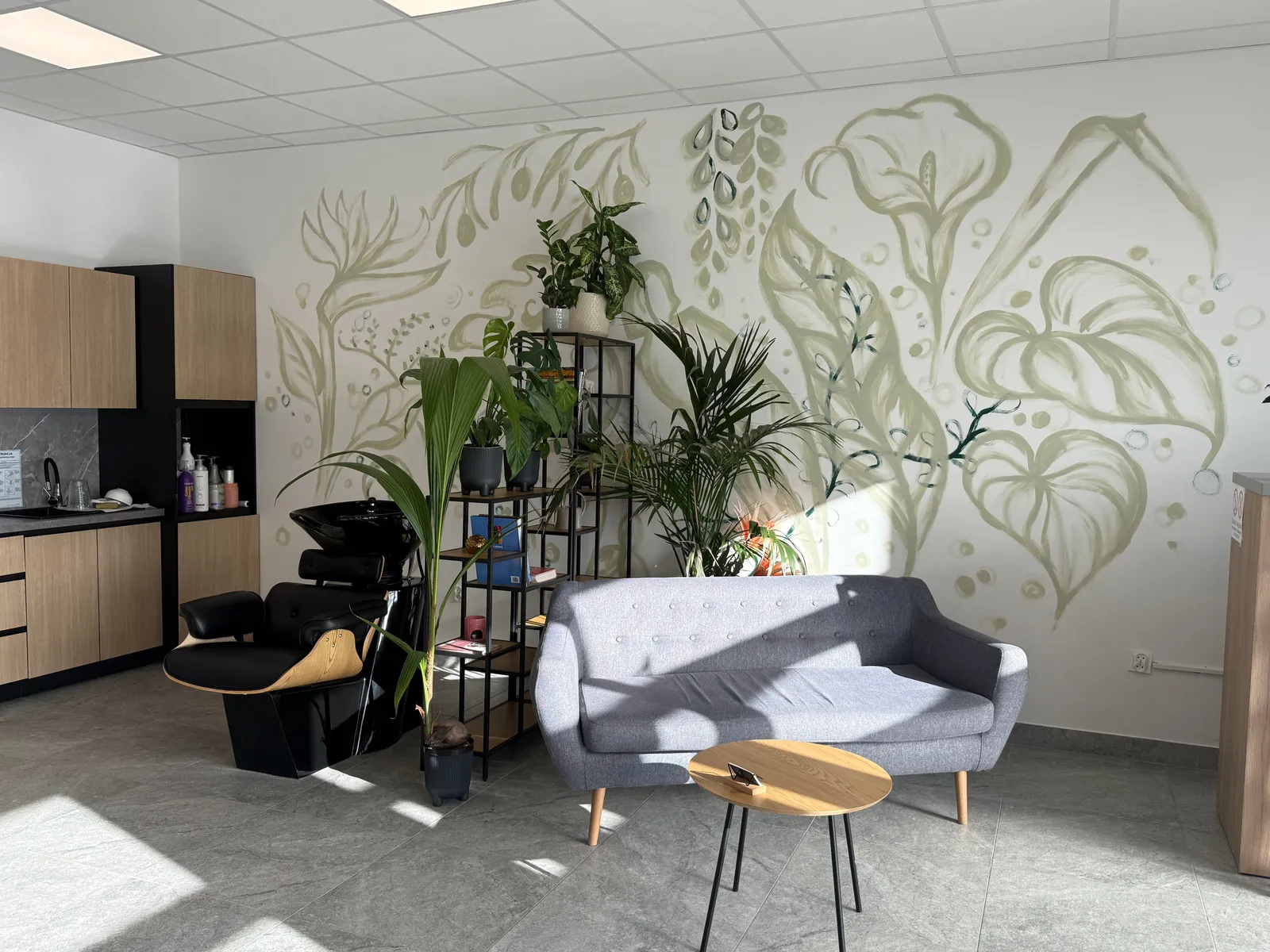 Wnętrze salonu AZ Beauty Loft: mural z zielonych liści, szara sofa, rośliny i drewniany stolik